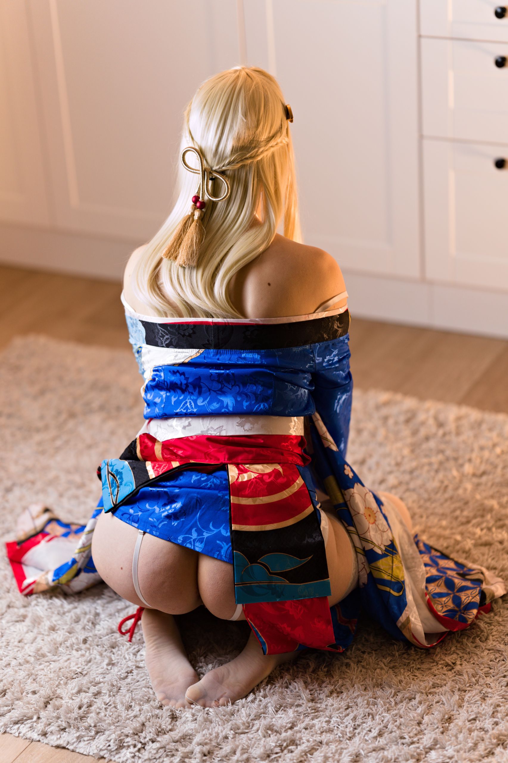 Shizuka Kawa cosplaying as Asuma Toki from Blue Archive dressed in a colorful kimono with white body harness underneath. Photo taken from above, as Shizuka sits on a carpet. Showcases her bare shoulders and partially exposed butt with prominent harness straps digging into it. Prominent hair accessory visible.
