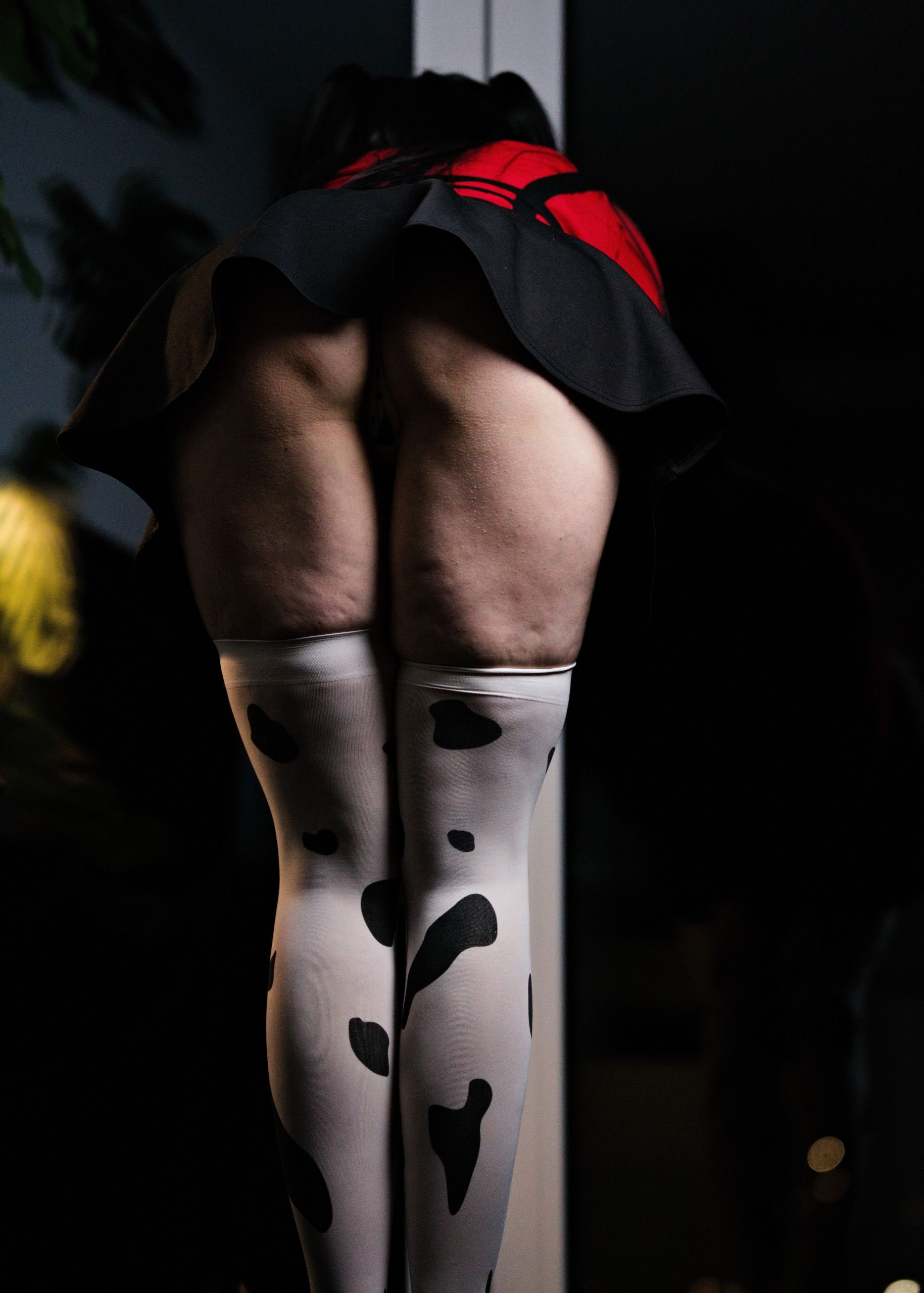 Shizuka Kawa cosplaying as Fuyuko Mayuzumi from Idolmaster wearing red shirt, black skirt with suspenders and cow-print stockings. Leaning forward and away from the viewer, allowing for an upskirt photo taken from below. Focus on legs in cow-print stockings, thick thighs and ass that's partially uncovered.