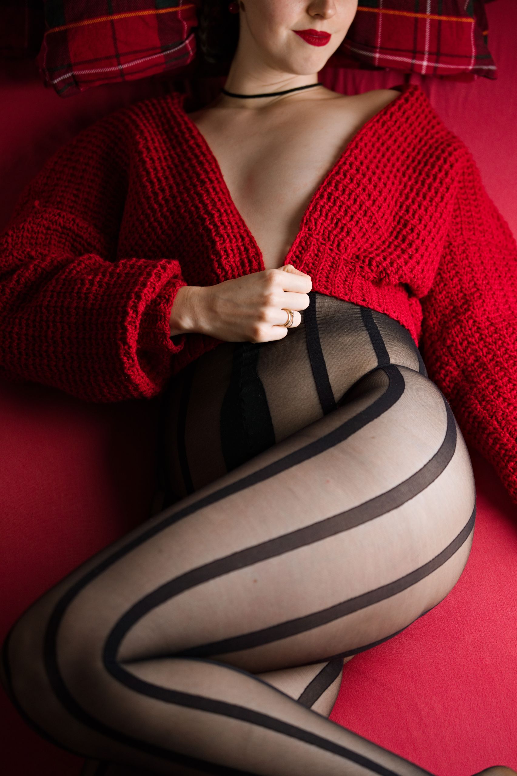 Shizuka Kawa dressed in a crop red knitted sweater with black bodystocking with vertical stripes underneath. Photo taken from above while she's lying down on a bed facing the viewer. Clevage visible but breasts are covered by sweater panels. Highlighted sheer bodystocking panels, micro thong and choker. Red lipstick visible. 