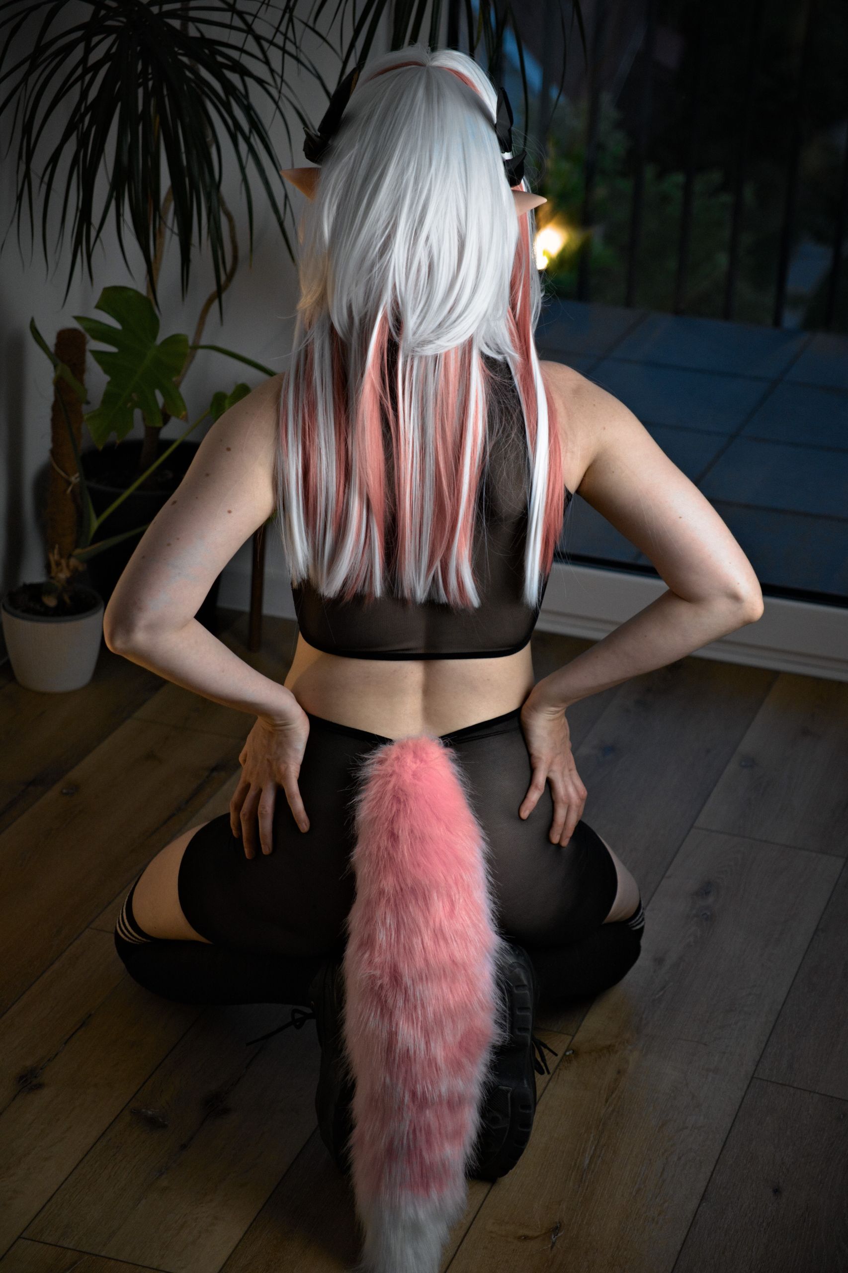 Shizuka Kawa cosplaying as Mi Fu in a black see-through mesh sporty outfit and white wig with pink highlights, posing while kneeling on the floor with with hands on hips and a pink fluffy tail and horns visible. Photo taken from behind.