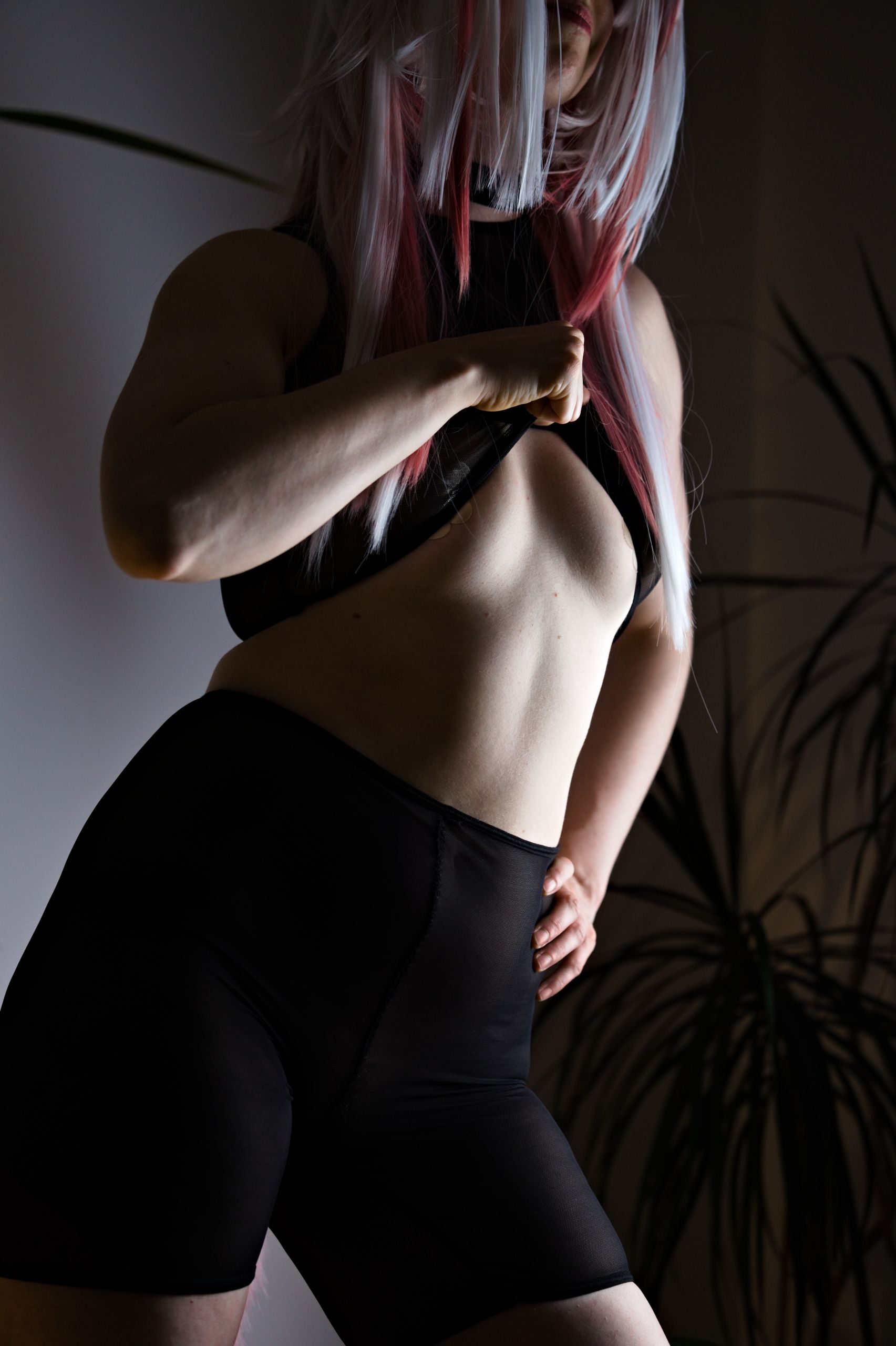 Shizuka Kawa cosplaying as Mi Fu wearing a black see-through mesh sporty outfit, posing with one hand on her hip and the other lifting her top to show bare cleavage and midriff.