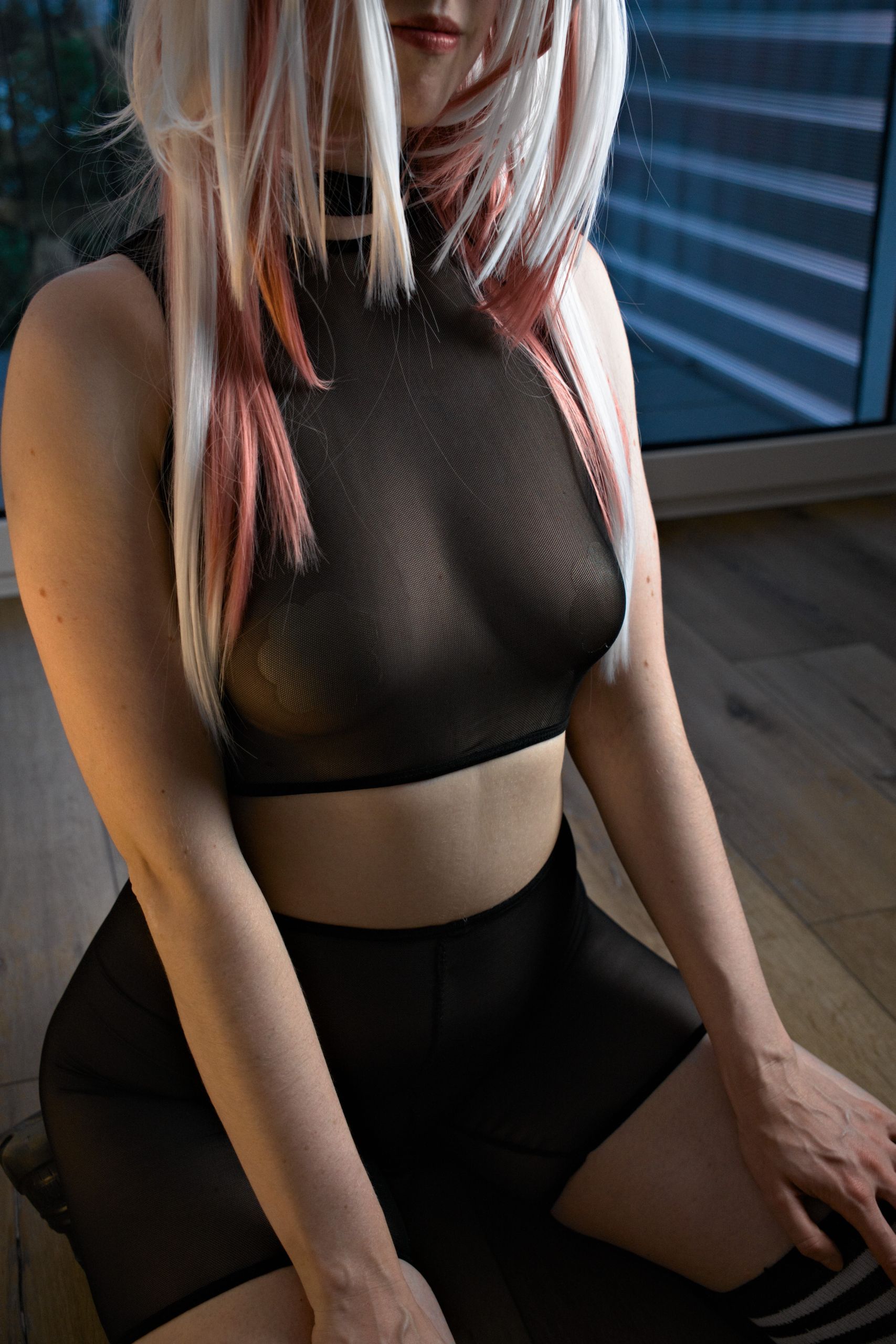 Shizuka Kawa cosplaying as Mi Fu from Arknights: Endfield dressed in a black see-through mesh sporty outfit consisting of a high-neck sleeveless crop top and matching shorts, sitting on a wooden floor with long white and pink highlighted hair visible.